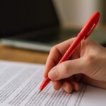 An image of a book editor holding red pen over blurred paperwork and laptop.